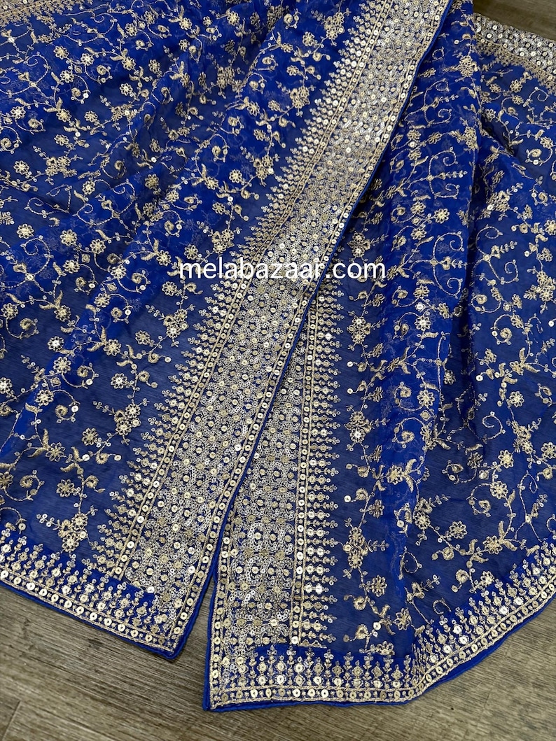 May include: Royal blue fabric with intricate gold floral embroidery and sequin detailing. The fabric is draped, showcasing the design and texture. The website melabazaar.com is visible on the fabric.