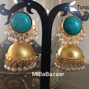 May include: A pair of gold-colored earrings with a turquoise stone and small white pearls. The earrings have a dome shape with a decorative edge and are displayed on a black stand. The word "Turquoise" is written in the top right corner and "MelaBazaar" is written at the bottom.