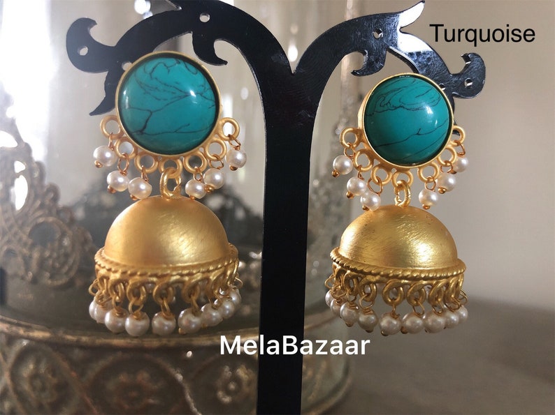 May include: A pair of gold-colored earrings with a turquoise stone and small white pearls. The earrings have a dome shape with a decorative edge and are displayed on a black stand. The word "Turquoise" is written in the top right corner and "MelaBazaar" is written at the bottom.
