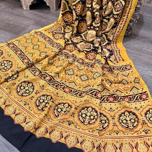 Puede incluir: Un textil plegado con un patrón detallado en negro, amarillo y rojo. El diseño incluye formas florales y geométricas. El textil tiene un borde negro y está drapeado sobre una superficie.