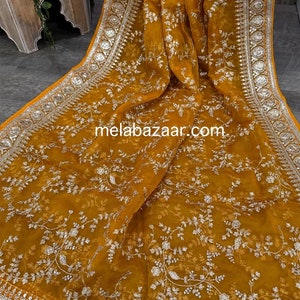 May include: A mustard yellow sheer scarf with a silver floral pattern and silver embroidered border. The scarf has a website address, "melabazar.com", printed on it.