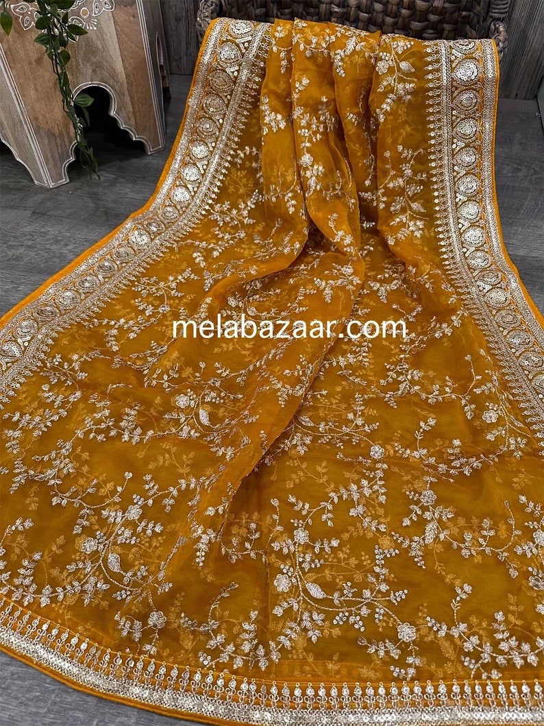 May include: A mustard yellow sheer scarf with a silver floral pattern and silver embroidered border. The scarf has a website address, "melabazar.com", printed on it.