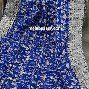 May include: Royal blue fabric with intricate gold floral embroidery and a silver sequined border. The fabric is draped, showcasing the detailed design and texture. The website melabazaar.com is visible on the fabric.