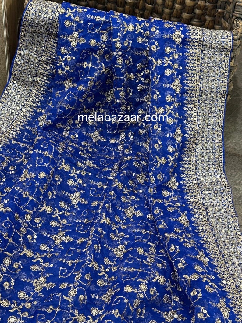 May include: Royal blue fabric with intricate gold floral embroidery and a silver sequined border. The fabric is draped, showcasing the detailed design and texture. The website melabazaar.com is visible on the fabric.