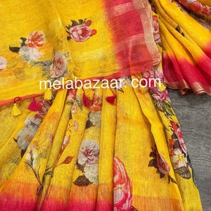 May include: Yellow linen saree with a pink border and floral print. The saree has a gradient effect with a darker yellow at the top and a lighter yellow at the bottom. The floral print is in shades of pink, white, and green.