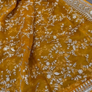 May include: A mustard yellow sheer fabric with a silver floral embroidery pattern. The fabric has a silver beaded border.