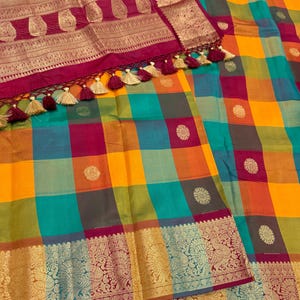 May include: A vibrant sari featuring a multi-colored checkered pattern in yellow, teal, and magenta. The fabric is adorned with small gold circular motifs and finished with a wide, ornate magenta border featuring gold metallic peacock designs.