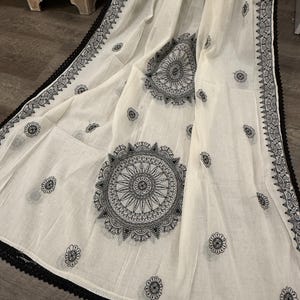 Pode incluir: Tecido branco com desenhos de mandala pretos e detalhes florais. As bordas s&atilde;o adornadas com uma borda de renda preta. O tecido &eacute; drapeado, mostrando os padr&otilde;es e texturas intrincados.