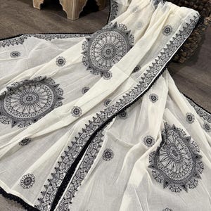 Pode incluir: Len&ccedil;o creme com intrincados padr&otilde;es de mandala pretos e detalhes florais. O len&ccedil;o apresenta uma borda de renda preta. O tecido &eacute; drapeado sobre uma superf&iacute;cie de madeira escura.