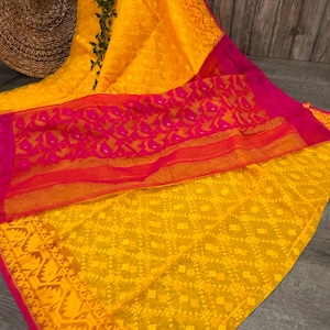 May include: A vibrant yellow textile with a repeating geometric pattern. A contrasting pink and orange sheer fabric is layered over the yellow, creating a colorful and textured effect. The edges are trimmed with a decorative border.
