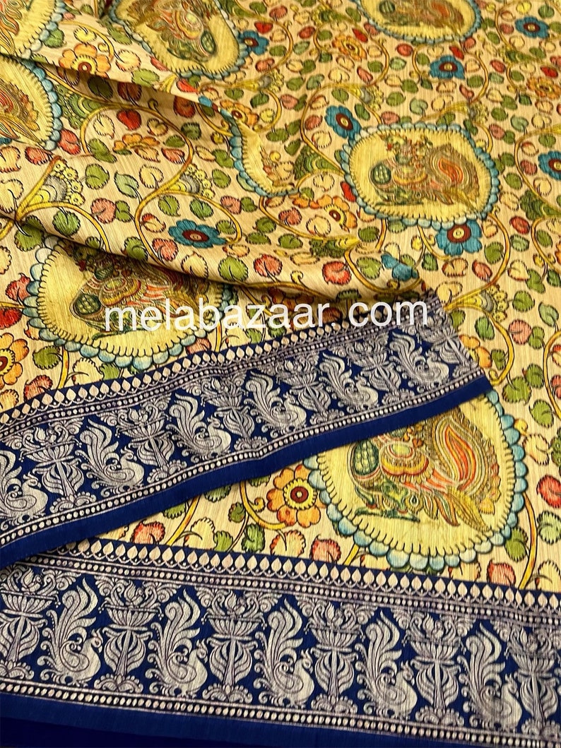 May include: A cream-colored fabric with a blue border and a floral pattern. The fabric features a repeating design of peacocks and flowers in shades of green, yellow, blue, and red. The fabric is called "melabazaar.com".