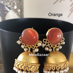 May include: A pair of orange and gold-colored earrings. The earrings have a large, round orange stone set in a gold frame, with a smaller gold bell-shaped bottom. Small white pearls hang from the gold frame and the bottom of the bell.