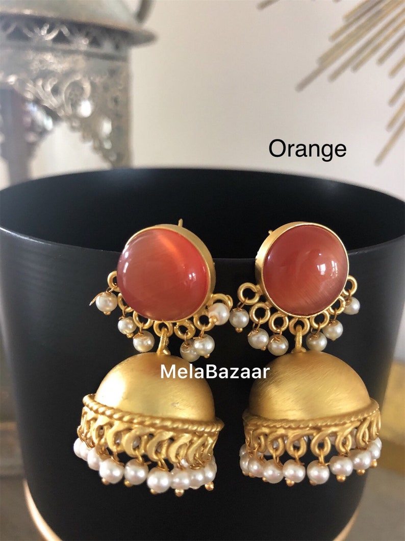 May include: A pair of orange and gold-colored earrings. The earrings have a large, round orange stone set in a gold frame, with a smaller gold bell-shaped bottom. Small white pearls hang from the gold frame and the bottom of the bell.