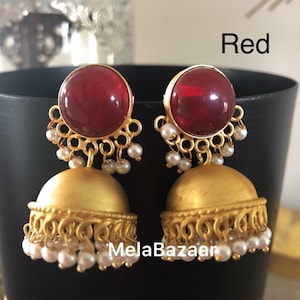 May include: A pair of gold-colored earrings with a red, rounded top and a bell-shaped bottom. The earrings are adorned with small white pearls and the word "Red" is visible in the image.
