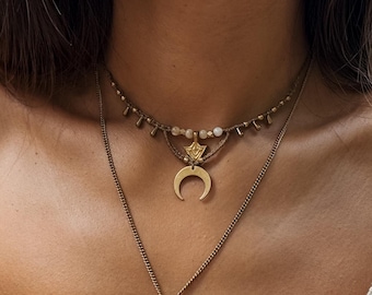 May include: A layered necklace set featuring three gold-toned chains. The top chain has small beads, the middle chain has a crescent moon pendant, and the bottom chain has a snake pendant. The necklaces are worn over a cream-colored top.