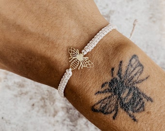 May include: A white string bracelet with a gold bee charm. The bracelet is on a person's wrist with a bee tattoo.