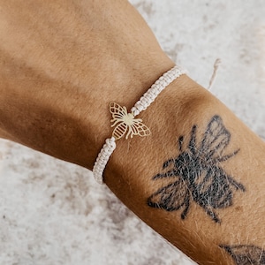 以下が含まれることがあります： 白い紐のブレスレットに金の蜂のチャームが付いている。ブレスレットは蜂のタトゥーのある人の腕につけられている。