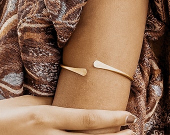 May include: A gold-toned metal arm cuff with a simple, minimalist design. The cuff is open on one side and has a slightly curved shape.