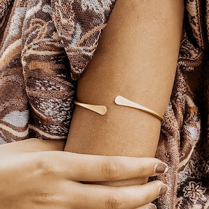 May include: A gold-toned metal arm cuff with a simple, minimalist design. The cuff is open on one side and has a slightly curved shape.