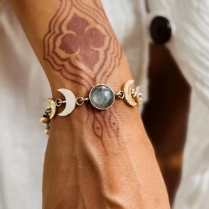 May include: A gold-toned bracelet featuring a central round grey stone flanked by two crescent moon charms. The bracelet is constructed with metal links connecting the moon and stone elements, creating a celestial-inspired design on a chain.