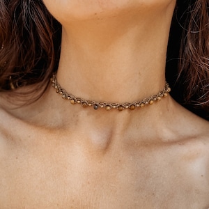 May include: A close-up of a woman wearing a gold beaded choker necklace. The necklace is made of small, round beads that are strung together on a thin chain. The woman is wearing a white shirt and has long, brown hair.