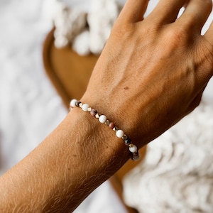 Könnte beinhalten: Ein Perlenarmband mit einem sich wiederholenden Muster aus glatten weißen Kugeln, braun marmorierten Kugeln und kleinen metallischen goldfarbenen Zwischenperlen, die auf einem elastischen Band aufgereiht sind.