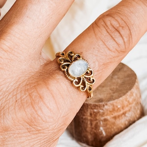 Può includere: Un anello color oro con una pietra di luna ovale e iridescente incastonata al centro. La fascia in metallo presenta un intricato design in filigrana traforata con motivi a volute che circondano l'incastonatura della gemma.