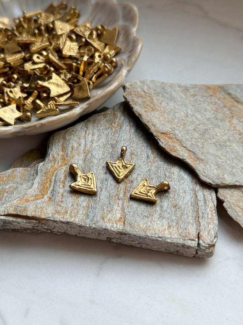 May include: Three gold-toned arrow-shaped pendants with a geometric design. The pendants have a loop at the top for attaching to a chain or cord.