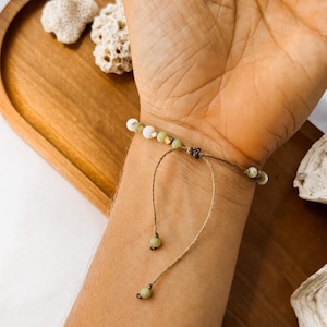 May include: A bracelet featuring small white and pale green beads accented with tiny gold-toned spacers. The bracelet is constructed with a thin brown cord that includes an adjustable sliding knot closure and two dangling cord ends finished with beads.