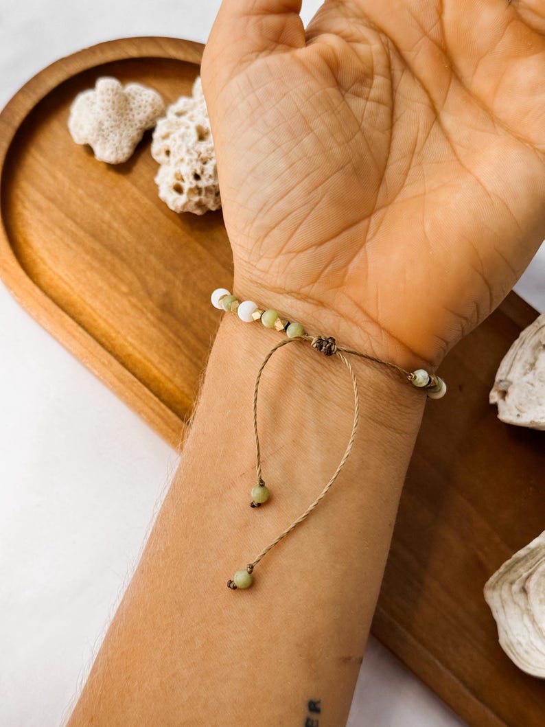 May include: A bracelet featuring small white and pale green beads accented with tiny gold-toned spacers. The bracelet is constructed with a thin brown cord that includes an adjustable sliding knot closure and two dangling cord ends finished with beads.