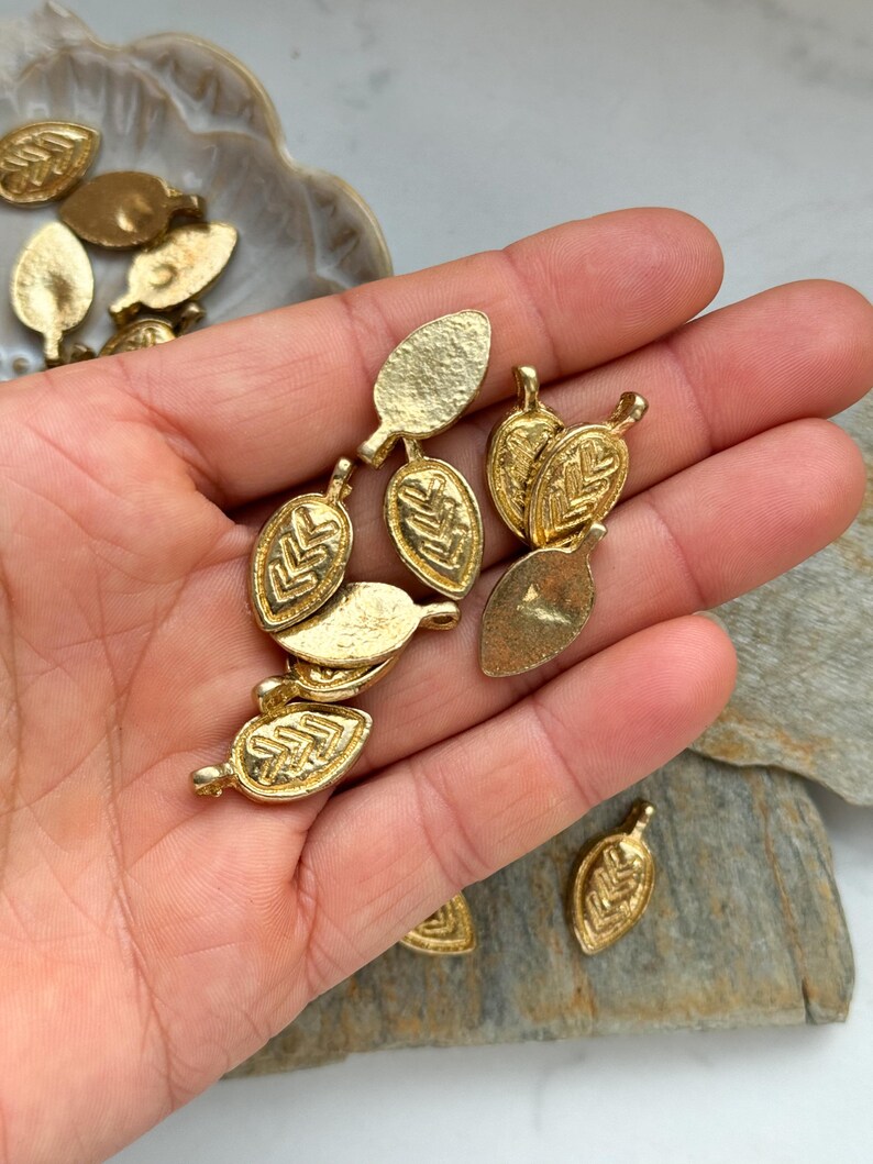 May include: A collection of gold-toned metal leaf charms with a textured surface and a geometric design. The charms have a hole at the top for attaching to jewelry or other crafts.