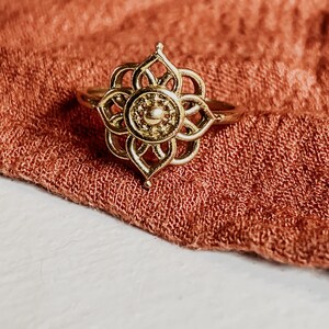K&ouml;nnte beinhalten: Ein goldfarbener Ring mit einem filigranen, kunstvollen Design, das an eine Blume erinnert. Der Ring liegt auf einem Hintergrund aus weichem, rostfarbenem Stoff.