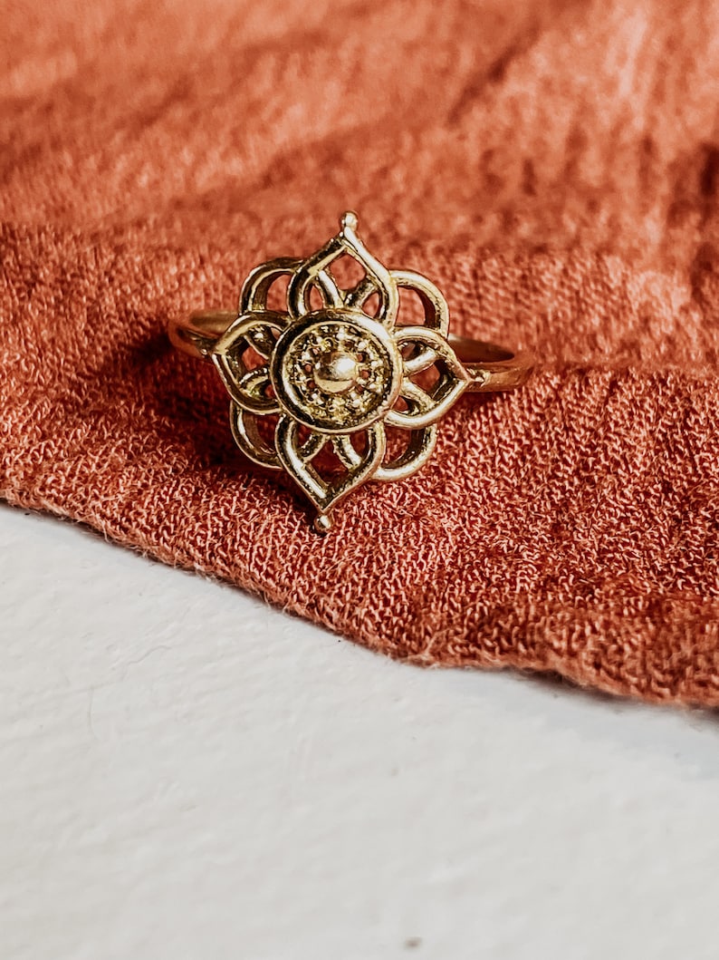 K&ouml;nnte beinhalten: Ein goldfarbener Ring mit einem filigranen, kunstvollen Design, das an eine Blume erinnert. Der Ring liegt auf einem Hintergrund aus weichem, rostfarbenem Stoff.