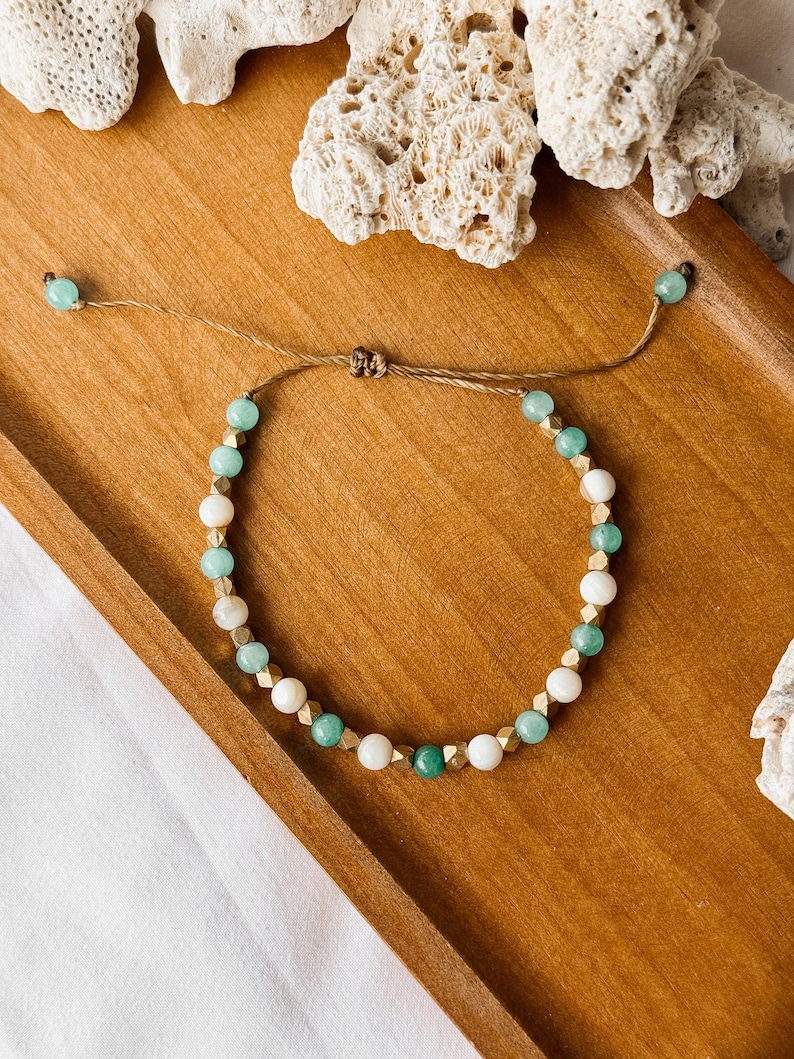 Puede incluir: Una pulsera de cuentas con cuentas redondas alternas de color verde menta y blanco, intercaladas con peque&ntilde;os espaciadores met&aacute;licos dorados facetados. La pulsera se asegura con un cord&oacute;n marr&oacute;n y tiene dos cuentas verde menta en los extremos.