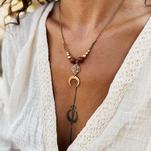 Könnte beinhalten: Eine goldfarbene Halskette mit einem Mond-Anhänger und einem floralen Design. Die Halskette hat braune und goldfarbene Perlen. Das Model trägt ein weißes Oberteil.