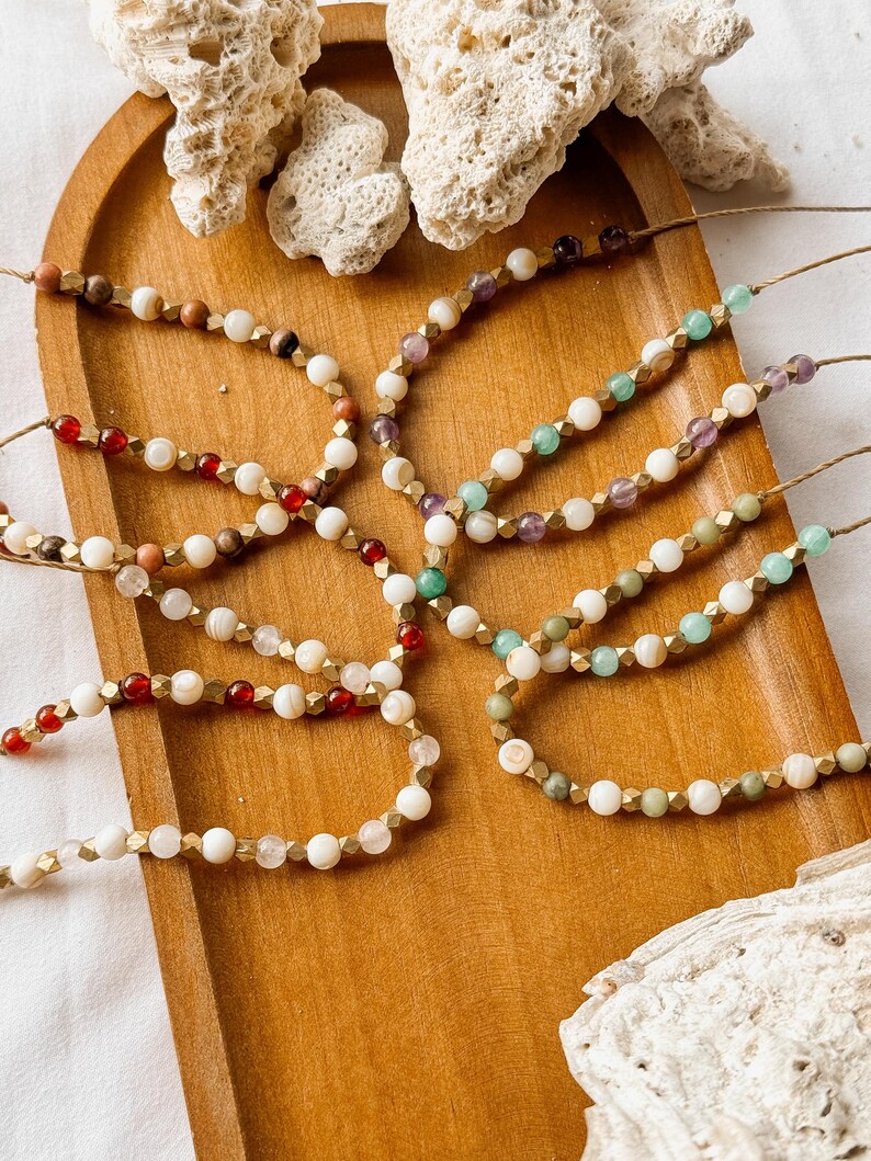 May include: A collection of six beaded bracelets arranged on a wooden tray. Each bracelet features a combination of white beads, metallic gold-toned faceted spacers, and various colored gemstone beads including red, green, and purple hues.
