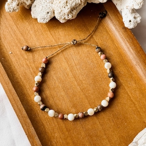 May include: A beaded bracelet featuring alternating round white, brown, and pink beads interspersed with small, faceted metallic gold-toned spacers. The bracelet is constructed with a thin brown cord and includes an adjustable sliding knot closure.