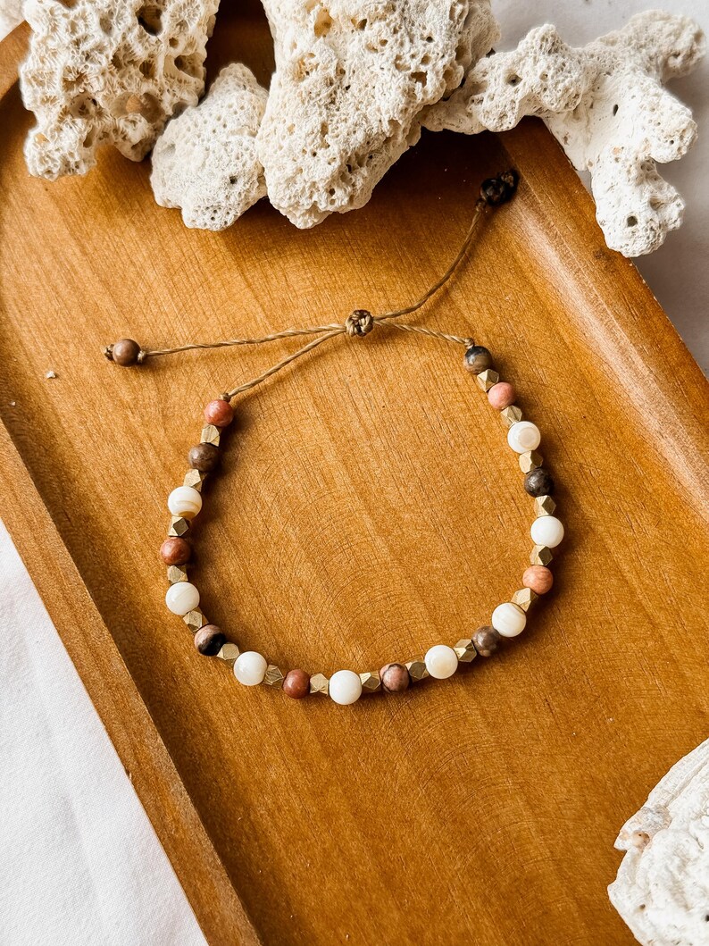May include: A beaded bracelet featuring alternating round white, brown, and pink beads interspersed with small, faceted metallic gold-toned spacers. The bracelet is constructed with a thin brown cord and includes an adjustable sliding knot closure.
