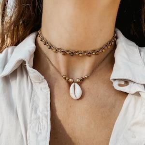 May include: A woman wearing a white shirt with a brown and gold beaded choker necklace and a shell pendant necklace.