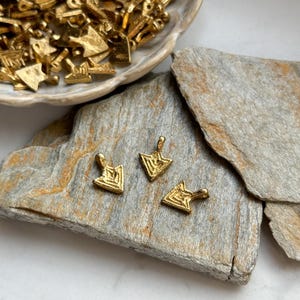 May include: Three gold arrow-shaped charms with a geometric pattern. The charms are on a light brown stone surface.