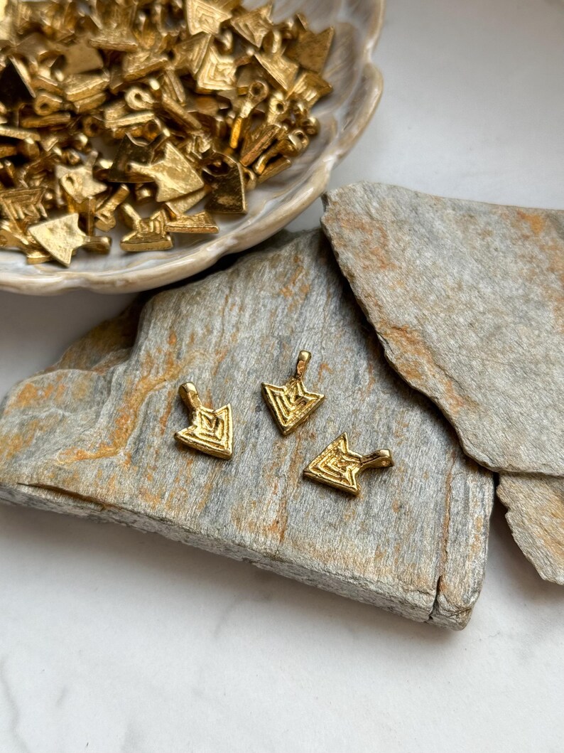 May include: Three gold arrow-shaped charms with a geometric pattern. The charms are on a light brown stone surface.