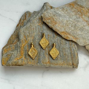 Puede incluir: Tres abalorios de metal de color dorado con un dise&ntilde;o geom&eacute;trico. Los abalorios tienen forma de diamante y una superficie texturizada.