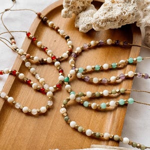 May include: A collection of adjustable bracelets made from natural twine, featuring a repeating pattern of white round beads, metallic gold-toned faceted spacers, and various colored gemstone beads including red, green, purple, and clear stones.