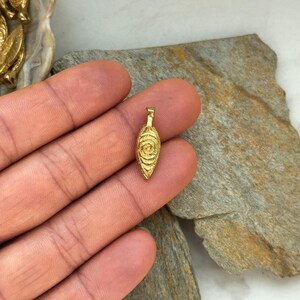 May include: A gold-colored pendant in the shape of an eye with a circular pattern. The pendant is on a hand.