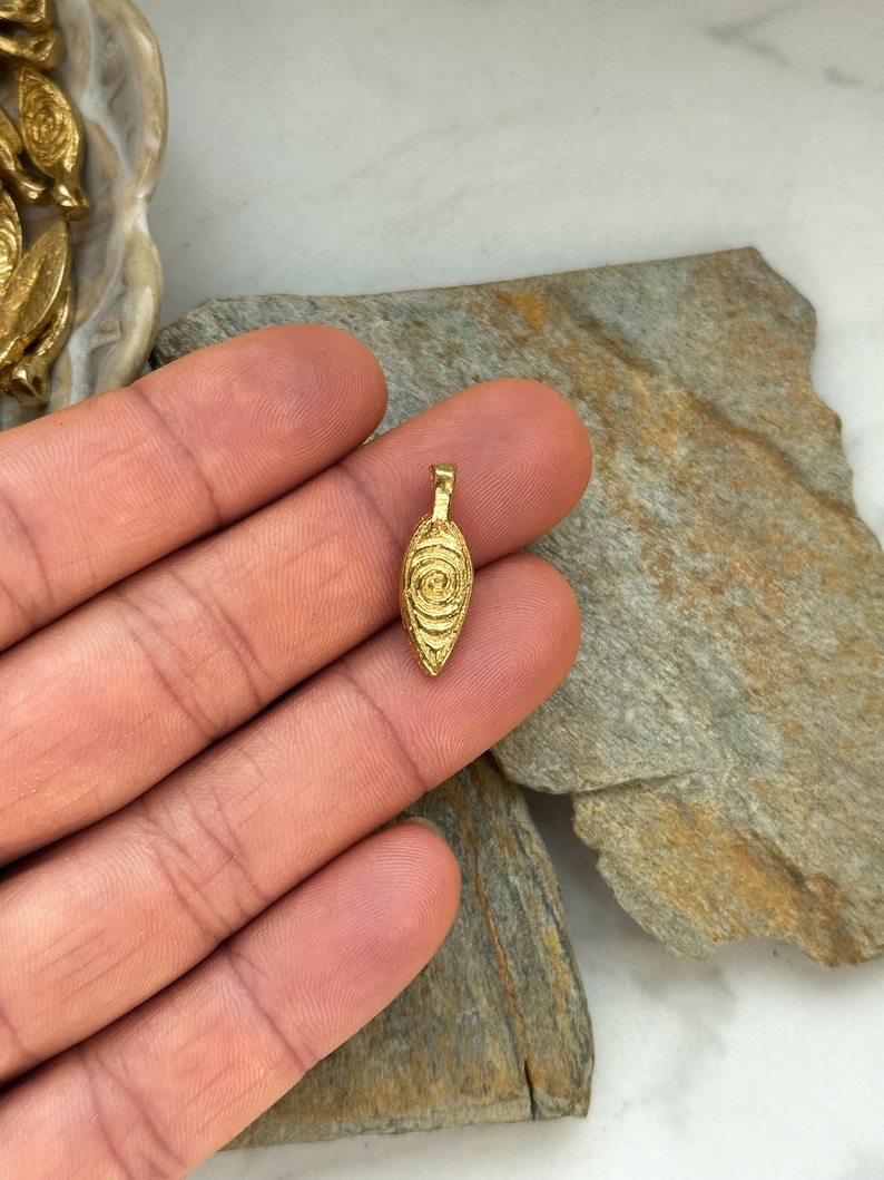 May include: A gold-colored pendant in the shape of an eye with a circular pattern. The pendant is on a hand.