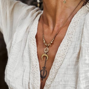 以下が含まれることがあります： 三日月ペンダントと幾何学的なチャームが付いたゴールドトーンのネックレス。 ネックレスは琥珀色のビーズが特徴で、お揃いのイヤリングと組み合わせています。 ジュエリーはクリーム色のブラウスを背景にしています。