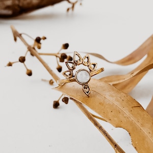 Puede incluir: Un anillo de oro con una piedra blanca engastada en un diseño delicado y ornamentado. El anillo está apoyado en una rama seca con hojas marrones.