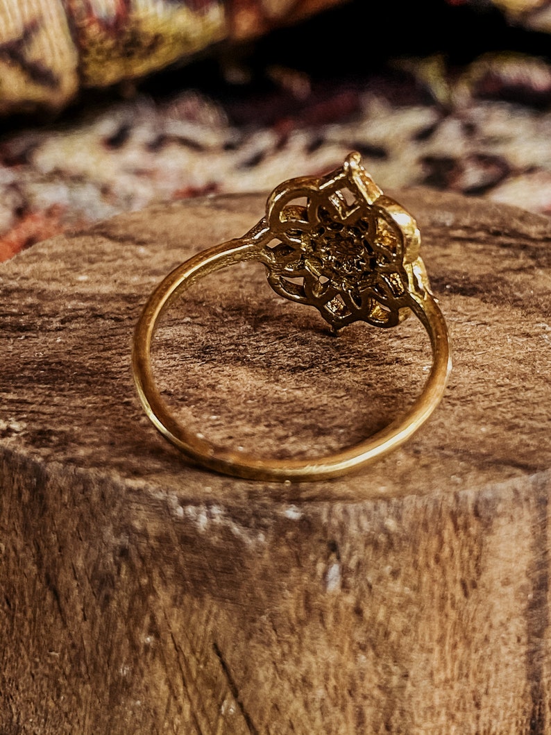 K&ouml;nnte beinhalten: Ein goldener Ring mit einem filigranen, komplizierten Design. Der Ring liegt auf einer Holzoberfl&auml;che.