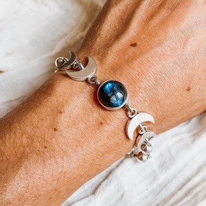 May include: A silver-toned bracelet featuring a central round cabochon stone with blue iridescent flashes, flanked by multiple crescent moon-shaped metal charms and small spherical beads connected by silver links in a chain design.