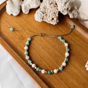 Puede incluir: Una pulsera de cuentas con una alternancia de cuentas redondas de color verde menta, cuentas blancas lisas y peque&ntilde;os espaciadores met&aacute;licos dorados. La pulsera est&aacute; ensartada en un cord&oacute;n marr&oacute;n fino con un nudo corredizo y dos cuentas verdes.